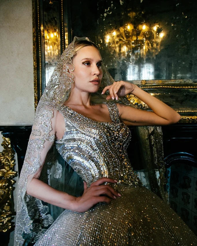 A crystal beaded gown with a matching embroidered hood and long sleeves, photographed seated beneath a chandelier.