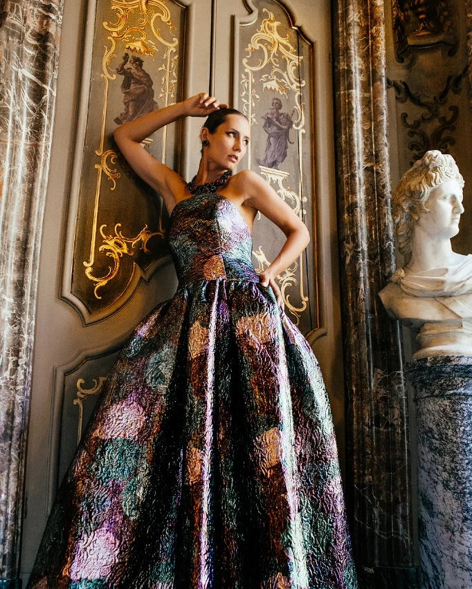An iridescent embroidered ball gown with a full skirt, photographed against gilded panelling beside a marble bust.