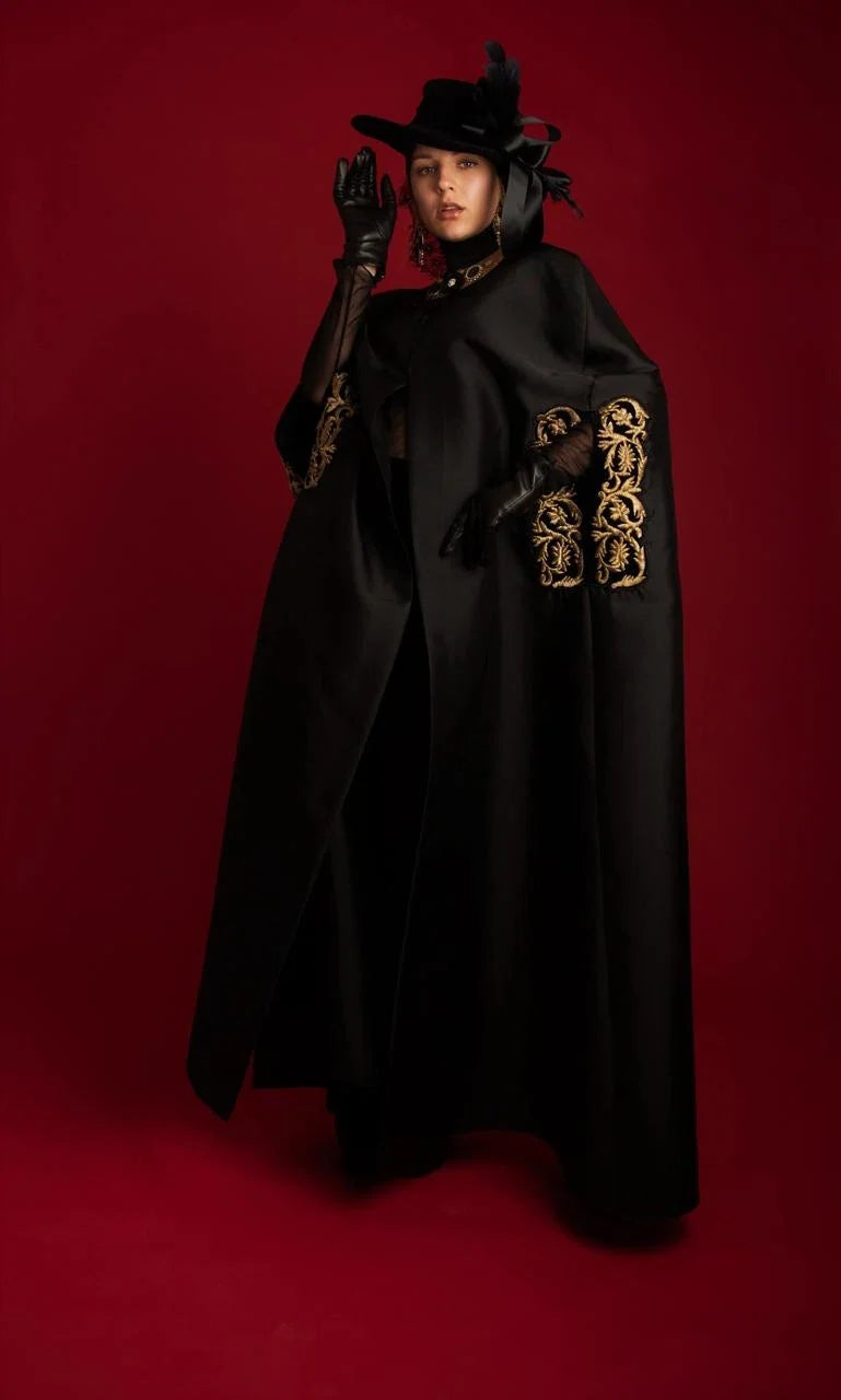 A black cape abaya with gold couched embroidery at the sleeves, photographed against red.