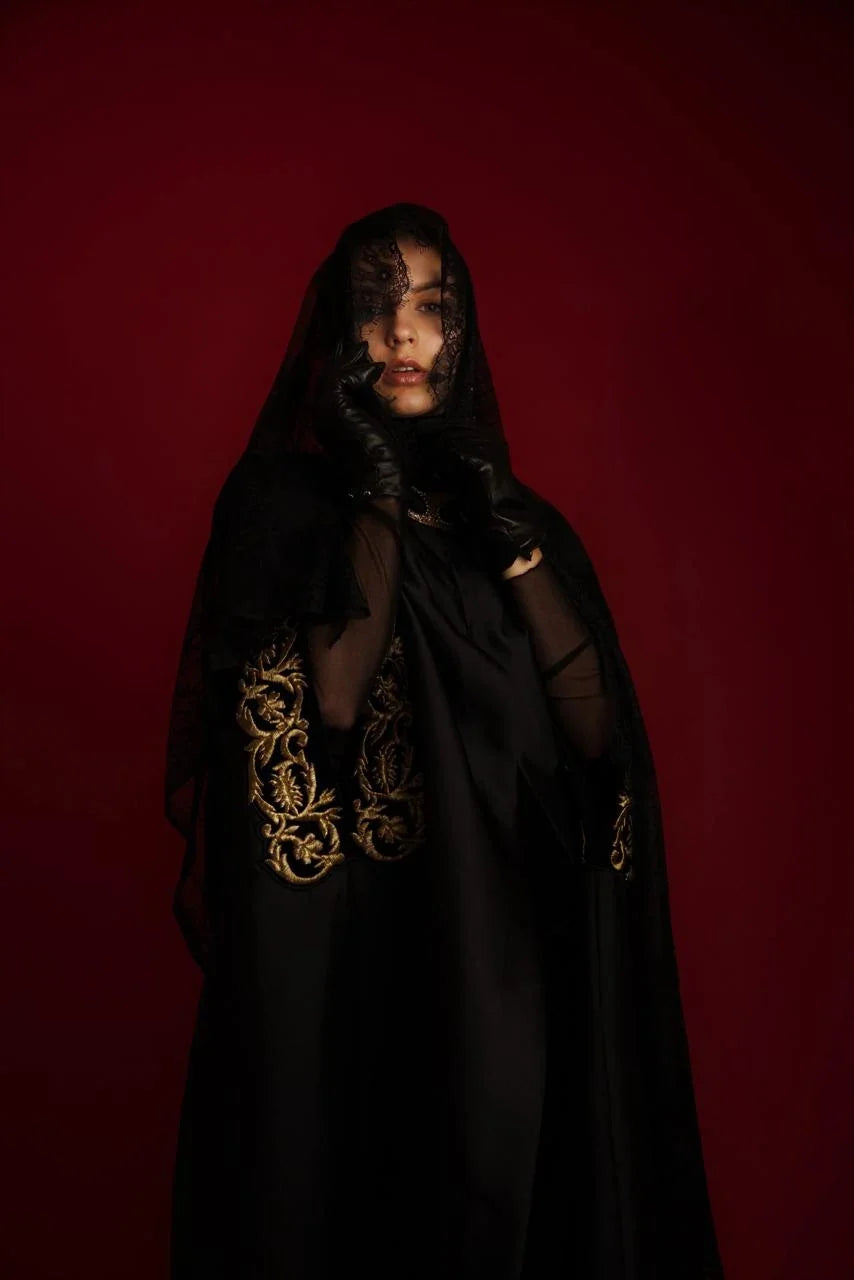 A black cape abaya with gold couched embroidery at the sleeves, photographed against red.