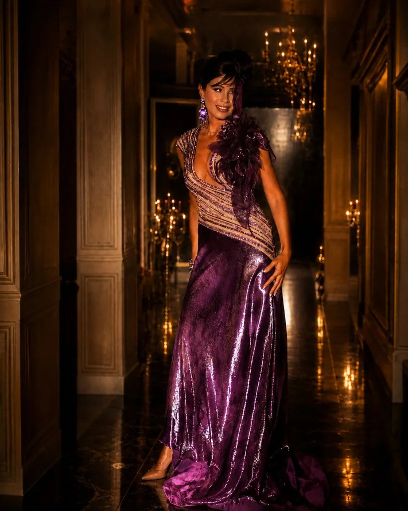 A plum metallic gown with a flower at the shoulder, photographed in a panelled corridor under chandeliers.