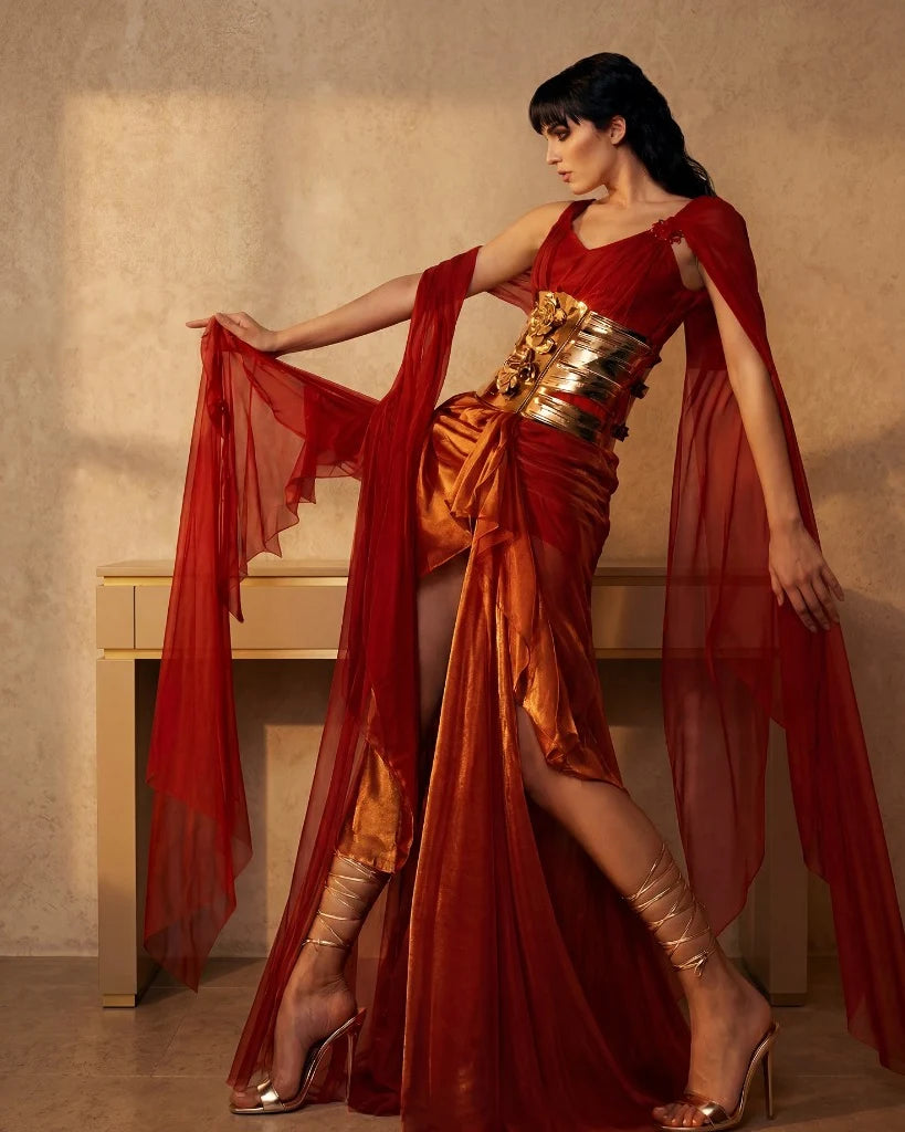 A red draped gown with a wide gold belt and chiffon panels held out at arm’s length.